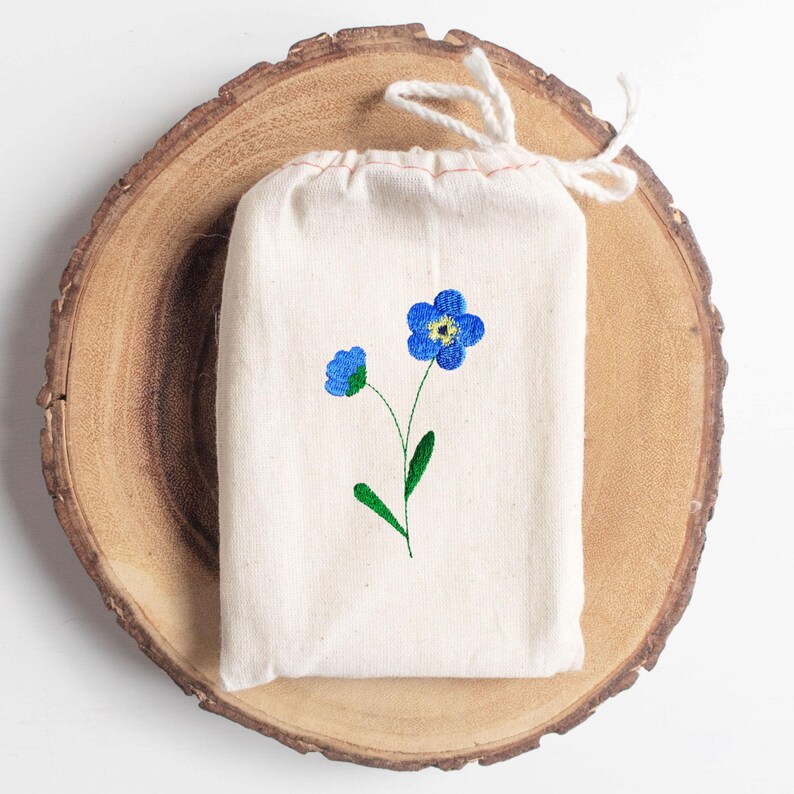 May include: A small, off-white drawstring bag with an embroidered blue forget-me-not flower design. The bag is made of a natural-colored fabric and sits on a wooden slice. The flower has a yellow center and green stem.