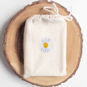 May include: A white cotton drawstring bag with a small embroidered daisy in the center. The daisy has a yellow center and white petals.
