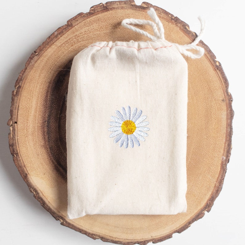 May include: A white cotton drawstring bag with a small embroidered daisy in the center. The daisy has a yellow center and white petals.