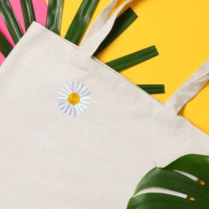 May include: A white canvas tote bag with a white daisy embroidered on the front. The bag has long handles and is lying on a yellow surface with green leaves.