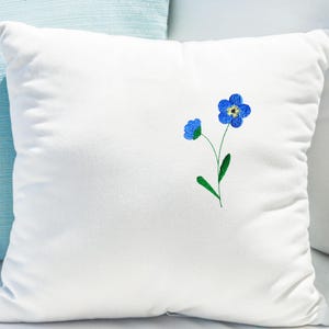May include: A square white pillow with a delicate embroidered design of blue forget-me-not flowers. The flowers have yellow centers and green stems and leaves. The pillow is made of a soft, plush material.