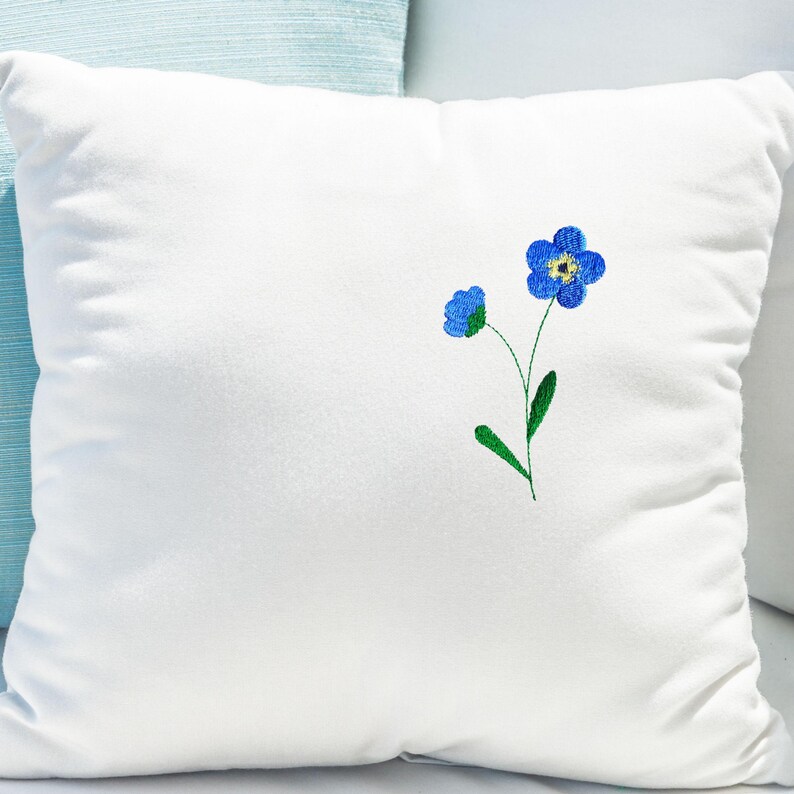 May include: A square white pillow with a delicate embroidered design of blue forget-me-not flowers. The flowers have yellow centers and green stems and leaves. The pillow is made of a soft, plush material.
