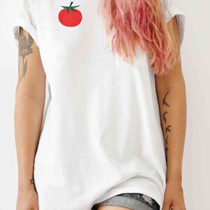 May include: A white t-shirt with a red tomato embroidered on the front. The model is wearing denim shorts and has pink hair.