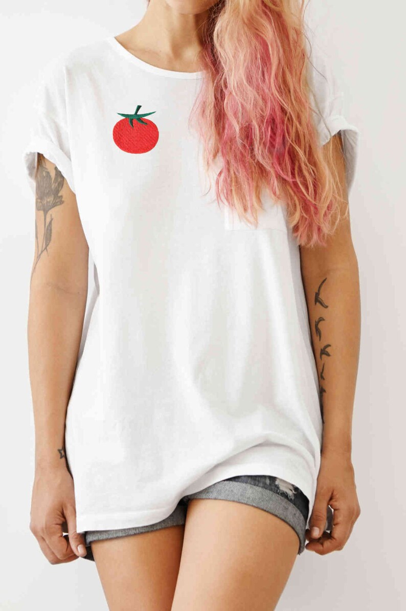 May include: A white t-shirt with a red tomato embroidered on the front. The model is wearing denim shorts and has pink hair.