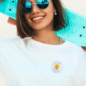 May include: A white t-shirt with a small embroidered daisy in the center. The daisy has a yellow center and white petals.