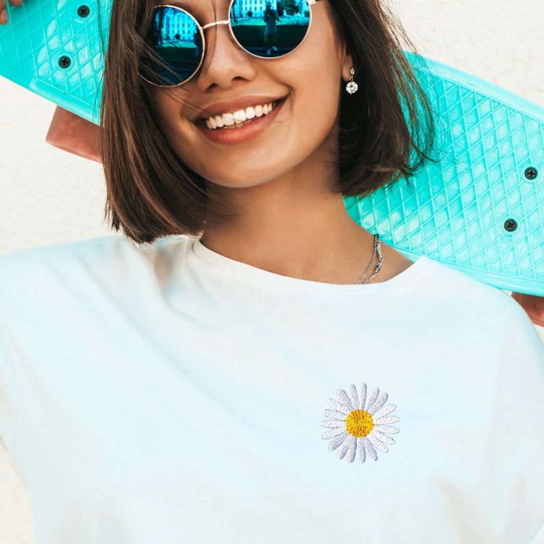 May include: A white t-shirt with a small embroidered daisy in the center. The daisy has a yellow center and white petals.