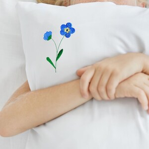 May include: White pillow with an embroidered blue forget-me-not flower design. The flower has a yellow center and green stem and leaves. The pillow is a decorative item.