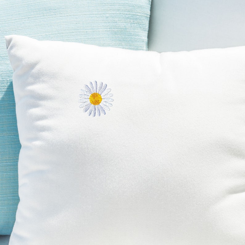 May include: A white pillow with a small embroidered daisy in the center. The daisy has a yellow center and white petals.