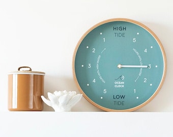 May include: A teal ocean clock with a wooden frame displays the tide level. A brown ceramic jar and a white decorative flower sit on a shelf above two pillows. One pillow is white with a brown zig-zag pattern.