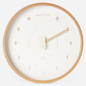 Op de afbeelding: Een witte houten wandklok met een lichtbruine houten lijst. De wijzerplaat van de klok heeft een maanfasenontwerp met de tekst "FULL MOON", "WANING MOON", "OCEAN CLOCK" en "WAXING MOON". De wijzers van de klok zijn van hout en wijzen naar de maanfasen.