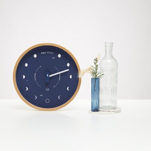 Op de afbeelding: Een ronde houten klok met een blauwe wijzerplaat en witte markeringen. De wijzerplaat van de klok heeft een maanfase-indicator en de tekst "FULL MOON".
