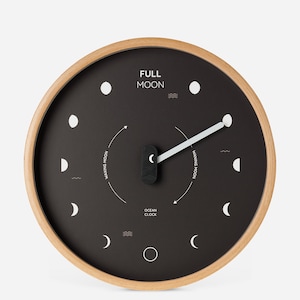 May include: Round wall clock with a black face and a light wood frame. The clock face displays the phases of the moon, with the words "FULL MOON" at the top. The clock hands are white, and the clock is labeled "OCEAN CLOCK".
