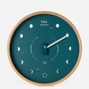Pode incluir: Um relógio circular azul-turquesa com moldura de madeira. O mostrador do relógio apresenta um ponteiro branco e um design de lua crescente. O texto "FULL MOON" e "OCEAN CLOCK" está impresso no mostrador do relógio.