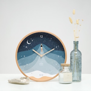Può includere: Un orologio da parete in legno con un design blu e bianco che presenta un'onda oceanica stilizzata e una persona che si tuffa in acqua. Il quadrante dell'orologio ha numeri romani e le parole "Ocean Clock" e "Six" stampate su di esso.