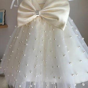 May include: A white tulle dress with a satin bow and pearl embellishments. The dress has short sleeves and a v-neckline.