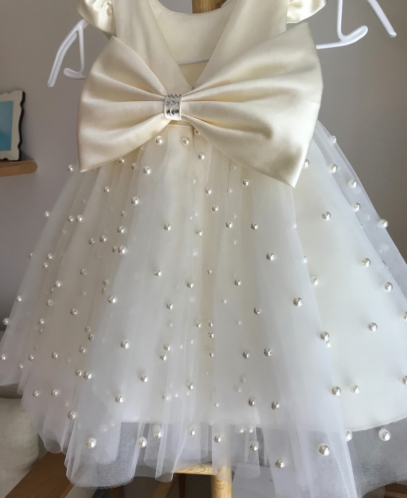 May include: A white tulle dress with a satin bow and pearl embellishments. The dress has short sleeves and a v-neckline.