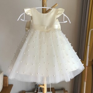 May include: A white tulle dress with a satin bodice and pearl embellishments. The dress has a bow at the waist and short sleeves.