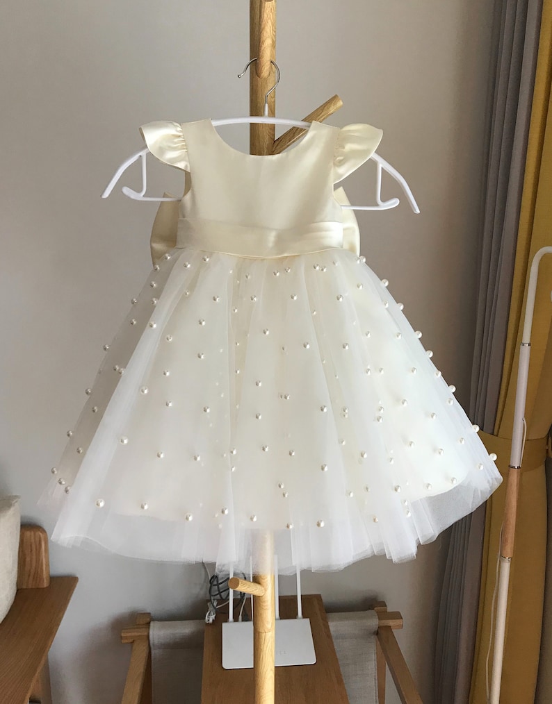 May include: A white tulle dress with a satin bodice and pearl embellishments. The dress has a bow at the waist and short sleeves.