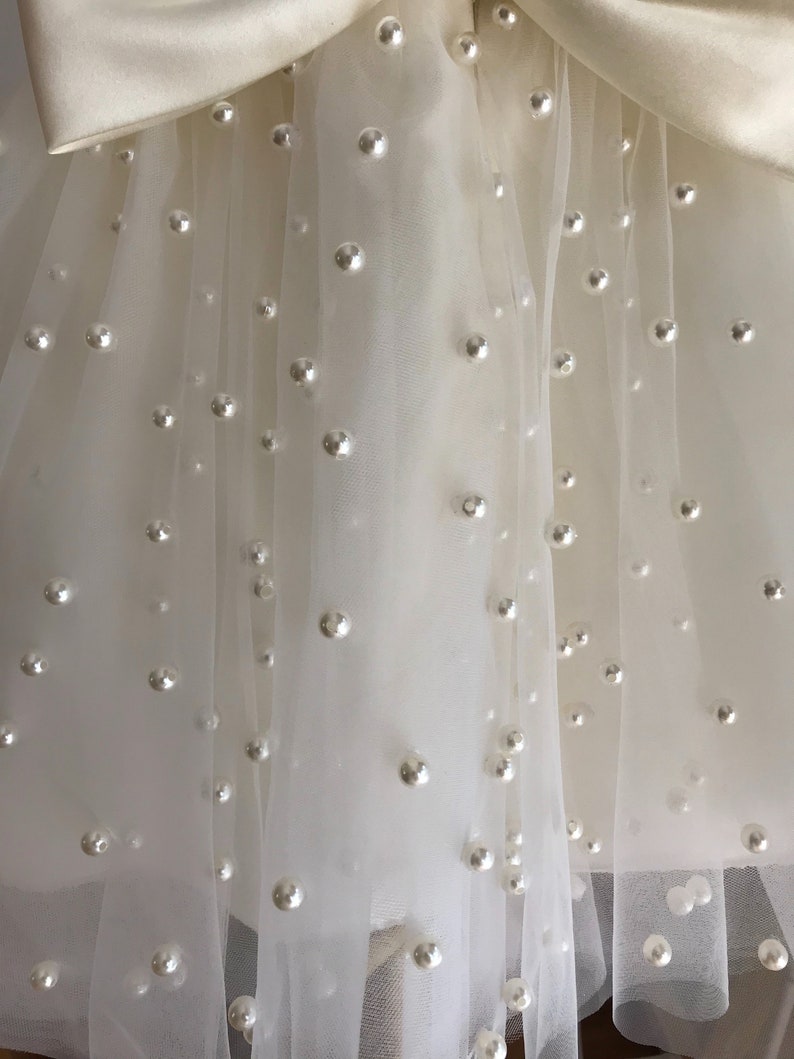May include: A white tulle skirt with a satin bow and pearl embellishments.