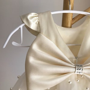 May include: A white tulle dress with a large satin bow and pearl embellishments. The dress has a sleeveless bodice and a full skirt.
