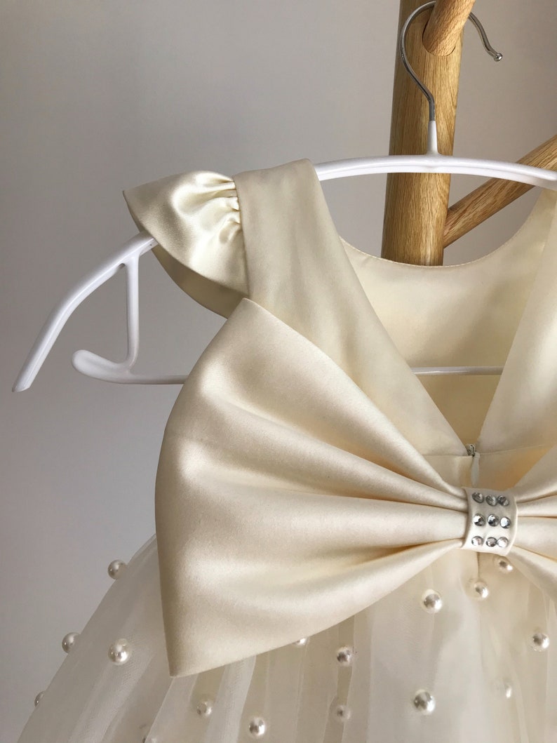 May include: A white tulle dress with a large satin bow and pearl embellishments. The dress has a sleeveless bodice and a full skirt.