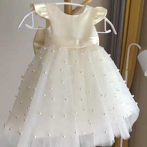 May include: A white tulle dress with pearl embellishments and a satin bodice. The dress has a bow at the waist and short, ruffled sleeves.