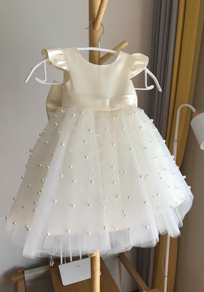 May include: A white tulle dress with pearl embellishments and a satin bodice. The dress has a bow at the waist and short, ruffled sleeves.