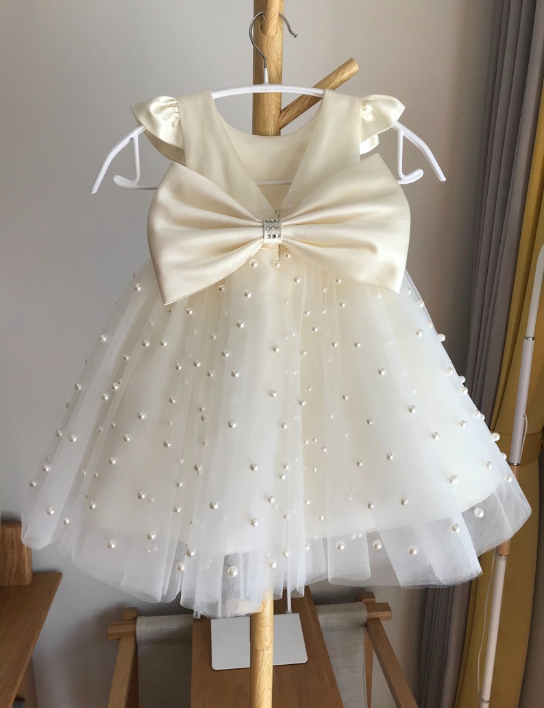 May include: A white tulle dress with a large satin bow and pearl embellishments. The dress has a V-neckline and short sleeves.