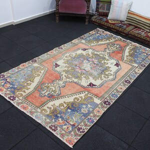 May include: A vintage Turkish rug with a cream background and a central medallion in shades of peach, blue, and green. The rug has a floral border with a repeating pattern of flowers and leaves.