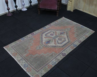 May include: A vintage Turkish rug with a light pink background and a geometric pattern in shades of beige, blue, and green. The rug has a fringed edge and is laid out on a black tiled floor.