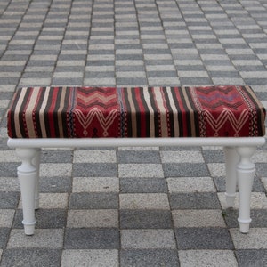 May include: White wooden bench with a red, brown and white patterned cushion. The bench is sitting on a grey tiled floor.