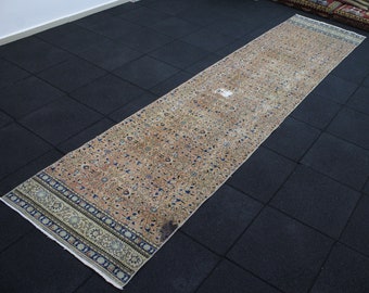 May include: A long, rectangular rug with an intricate floral pattern in shades of beige, blue, and brown. The rug has a distressed, vintage appearance, with a detailed border design. The rug is placed on a dark, tiled floor.