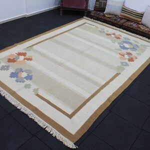 Puede incluir: Una alfombra de color crema con un borde marr&oacute;n y un patr&oacute;n floral en tonos azules, naranjas y verdes. La alfombra tiene flecos en los bordes.