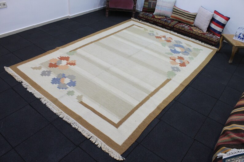 Puede incluir: Una alfombra de color crema con un borde marr&oacute;n y un patr&oacute;n floral en tonos azules, naranjas y verdes. La alfombra tiene flecos en los bordes.