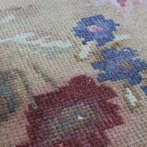 May include: Close-up of a beige rug with a floral pattern. The rug features flowers in shades of red, pink, and blue. The rug has a textured surface and a vintage aesthetic.