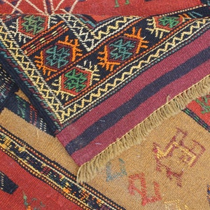 May include: A close-up of a colorful woven rug with intricate geometric patterns in shades of red, black, green, yellow, and brown. The rug has a fringe border and is made of wool.