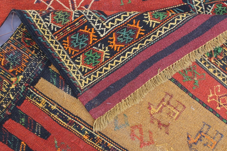 May include: A close-up of a colorful woven rug with intricate geometric patterns in shades of red, black, green, yellow, and brown. The rug has a fringe border and is made of wool.