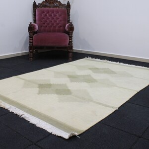 May include: A white rug with a diamond pattern in a light green color. The rug has a fringe border and is laid out on a black tiled floor.