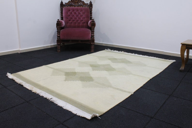 May include: A white rug with a diamond pattern in a light green color. The rug has a fringe border and is laid out on a black tiled floor.