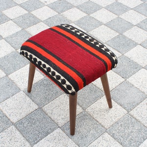 May include: A red, black, and orange striped ottoman with a wooden frame. The ottoman is sitting on a checkered tile floor.
