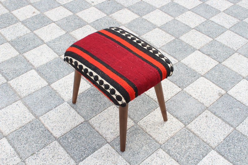 May include: A red, black, and orange striped ottoman with a wooden frame. The ottoman is sitting on a checkered tile floor.