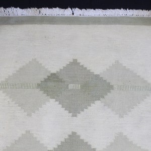May include: A white rug with a geometric pattern in shades of gray. The rug has a fringe border.