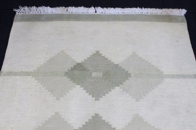 May include: A white rug with a geometric pattern in shades of gray. The rug has a fringe border.