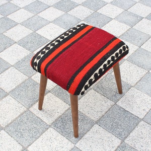 May include: A red, black, and white striped ottoman with wooden legs. The ottoman is sitting on a checkered floor.