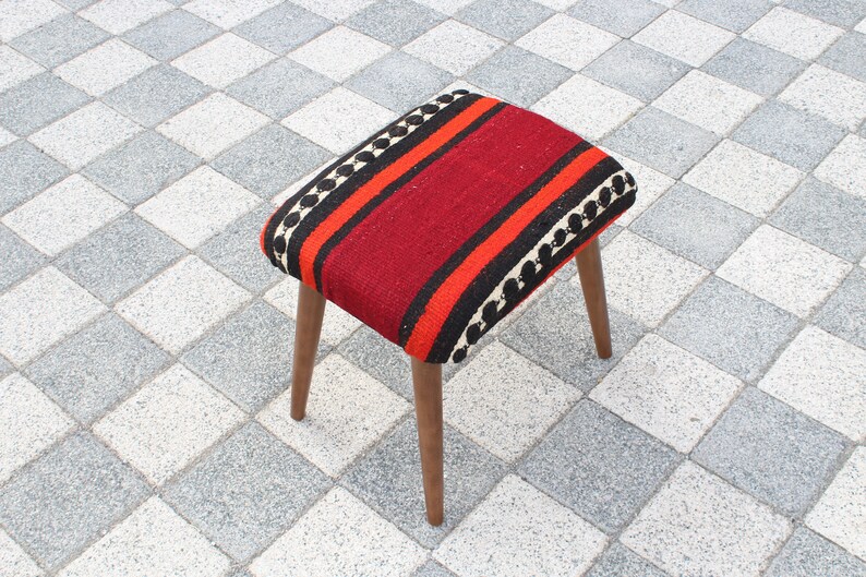 May include: A red, black, and white striped ottoman with wooden legs. The ottoman is sitting on a checkered floor.