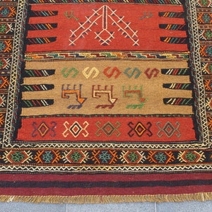 May include: A traditional rug with a red, brown, and black color scheme. The rug features intricate geometric patterns and a fringe border.