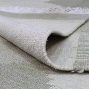 May include: Close-up of a white and light green woven rug with a fringe edge. The rug has a subtle pattern of stripes and geometric shapes.