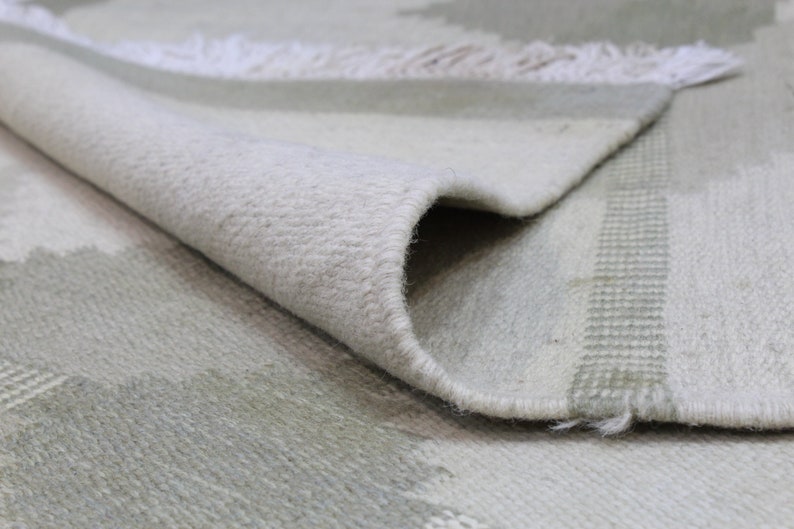 May include: Close-up of a white and light green woven rug with a fringe edge. The rug has a subtle pattern of stripes and geometric shapes.