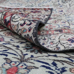 May include: A close-up of a cream-colored rug with a floral pattern in shades of blue, red, and brown. The rug is folded over, revealing the underside, which is a darker gray color.