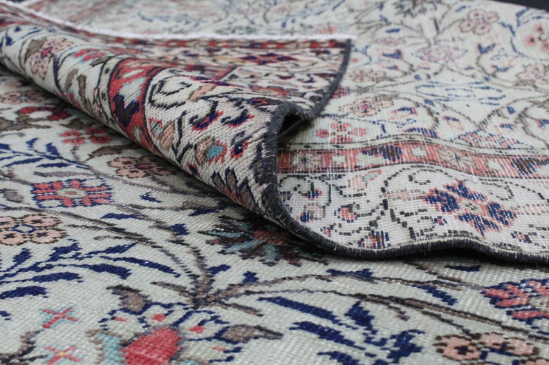 May include: A close-up of a cream-colored rug with a floral pattern in shades of blue, red, and brown. The rug is folded over, revealing the underside, which is a darker gray color.